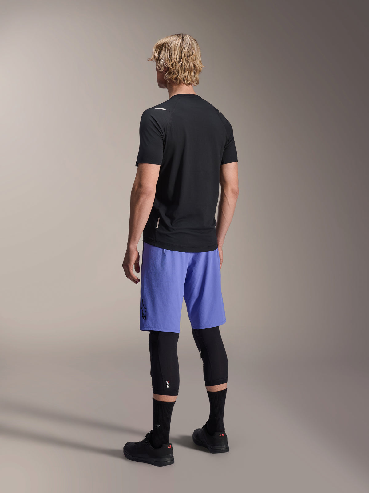 Alpinestars A-Aria Switch Jersey, jersey de MTB de manga corta, negro, vista trasera en modelo con pantalones cortos azules y rodilleras negras, equipamiento de ciclismo de alto rendimiento para trail
