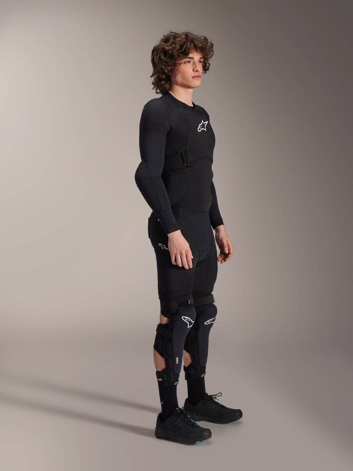 Chaqueta de protección Alpinestars A-Impact Plasma - Manga Larga, negra, usada por un modelo con equipo de protección a juego que incluye rodilleras y shorts, con logotipos blancos de Astars, construcción de malla transpirable y diseño estilizado para ciclismo de montaña