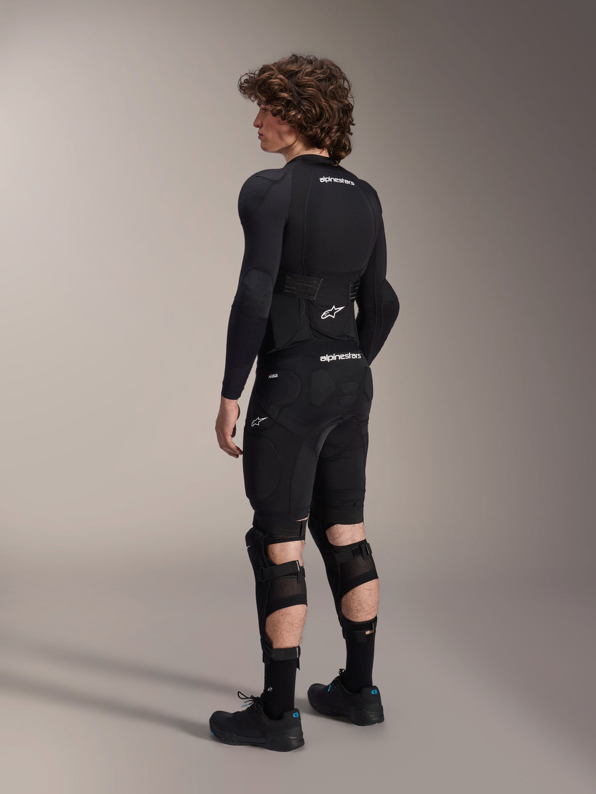 Modelo vistiendo los protectores de rodilla y espinilla Alpinestars A-Impact Plasma Pro desde atrás, en negro y blanco, mostrando el cierre de triple correa y los paneles de malla ventilada, combinados con un equipo completo de protección para bicicleta de montaña en color negro que incluye capa base de manga larga y pantalones cortos acolchados.