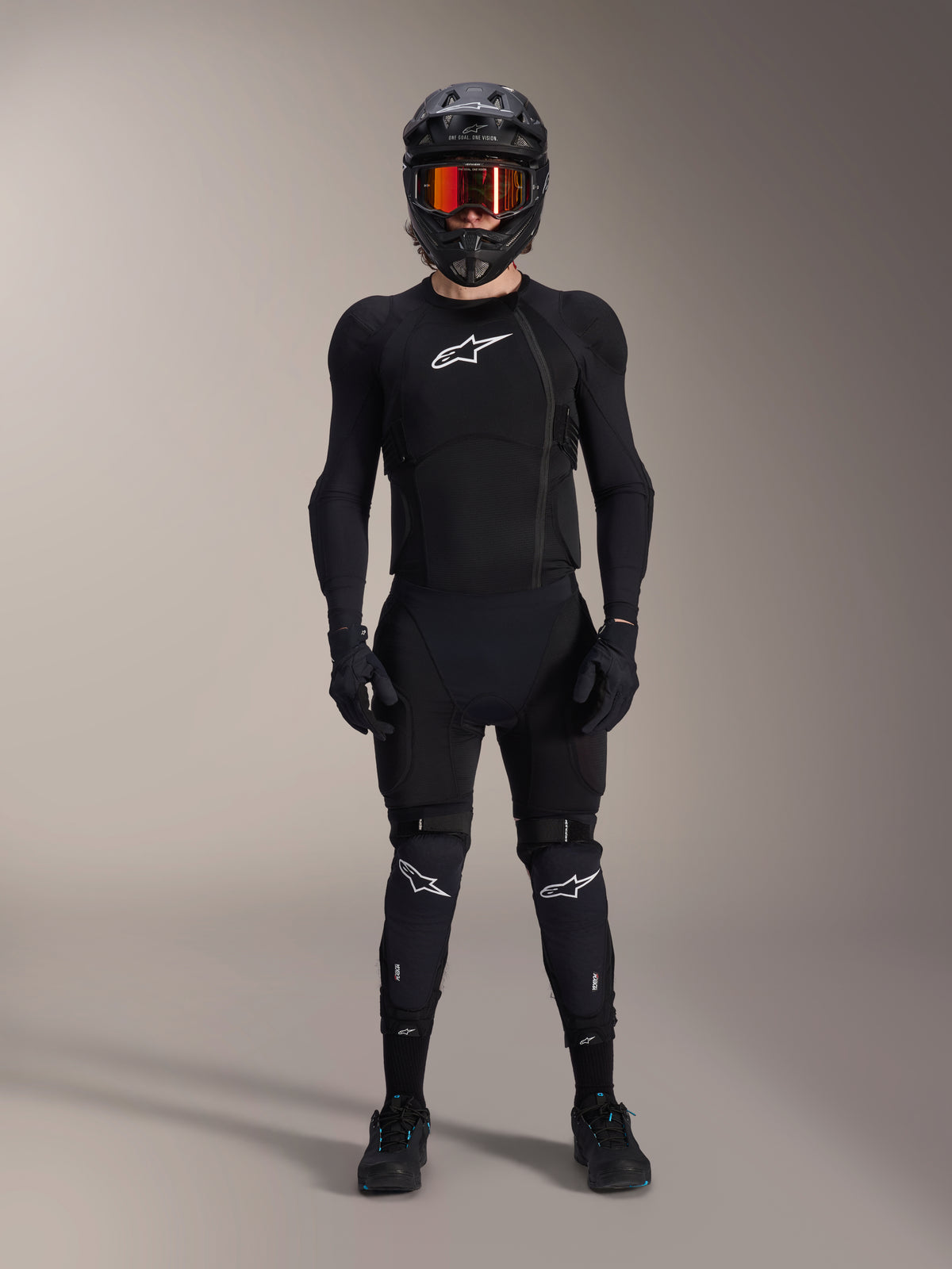 Ciclista de montaña con equipamiento de protección integral, chaqueta de protección Alpinestars A-Impact Plasma en negro y blanco, con logotipo blanco en el pecho y tejido de malla, acompañada de casco negro, gafas de lentes rojas y rodilleras, conjunto de armadura de MTB coordinado de alto rendimiento.