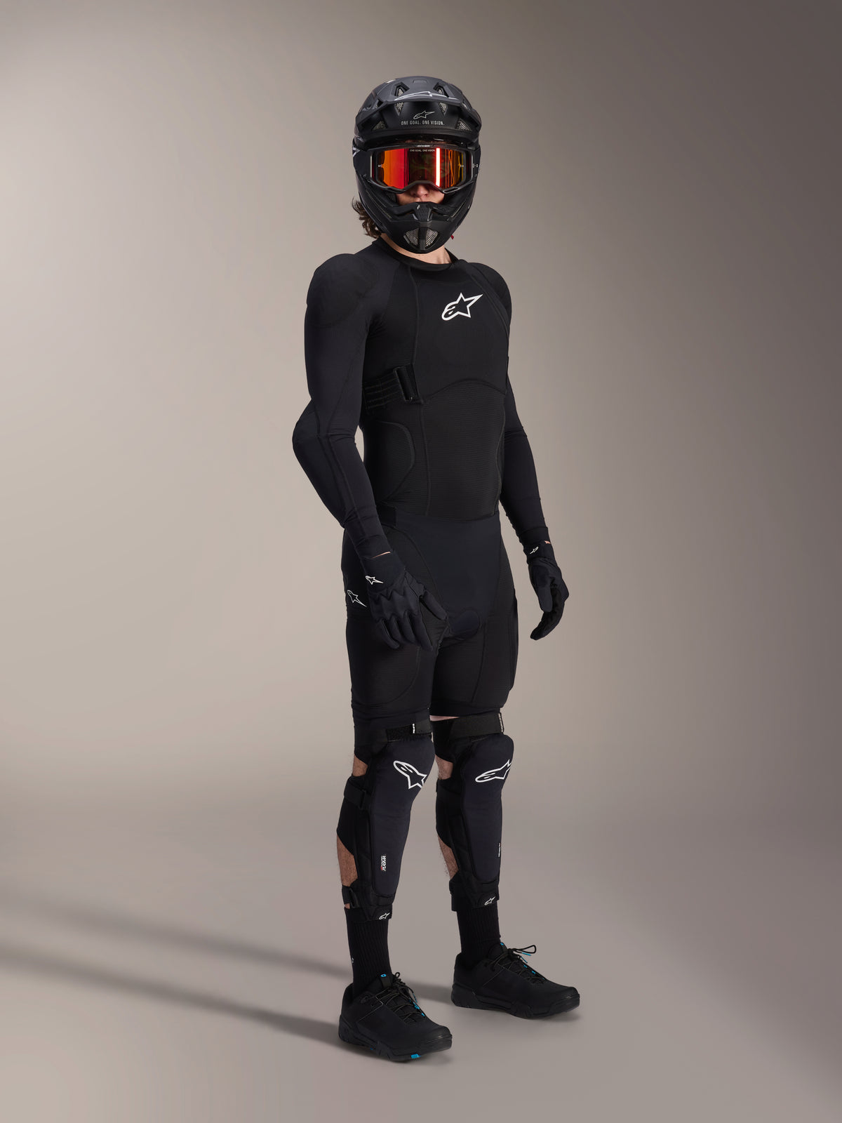 Protectores de rodilla y espinilla Alpinestars A-Impact Plasma Pro, en negro y blanco, usados por un ciclista con un equipamiento protector completo para bicicleta de montaña, que incluye una prenda superior blindada de manga larga negra, guantes negros y un casco integral con gafas naranjas.