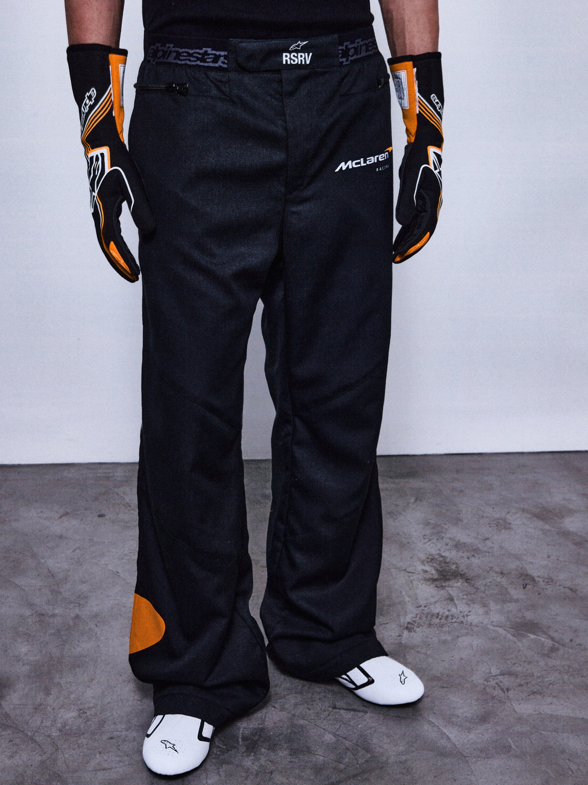 Alpinestars RSRV Arena Suit Pants, pantalones de carreras negros con parches naranjas, logos de McLaren y Alpinestars, persona usando guantes negros y naranjas, zapatos de carreras blancos con detalles negros