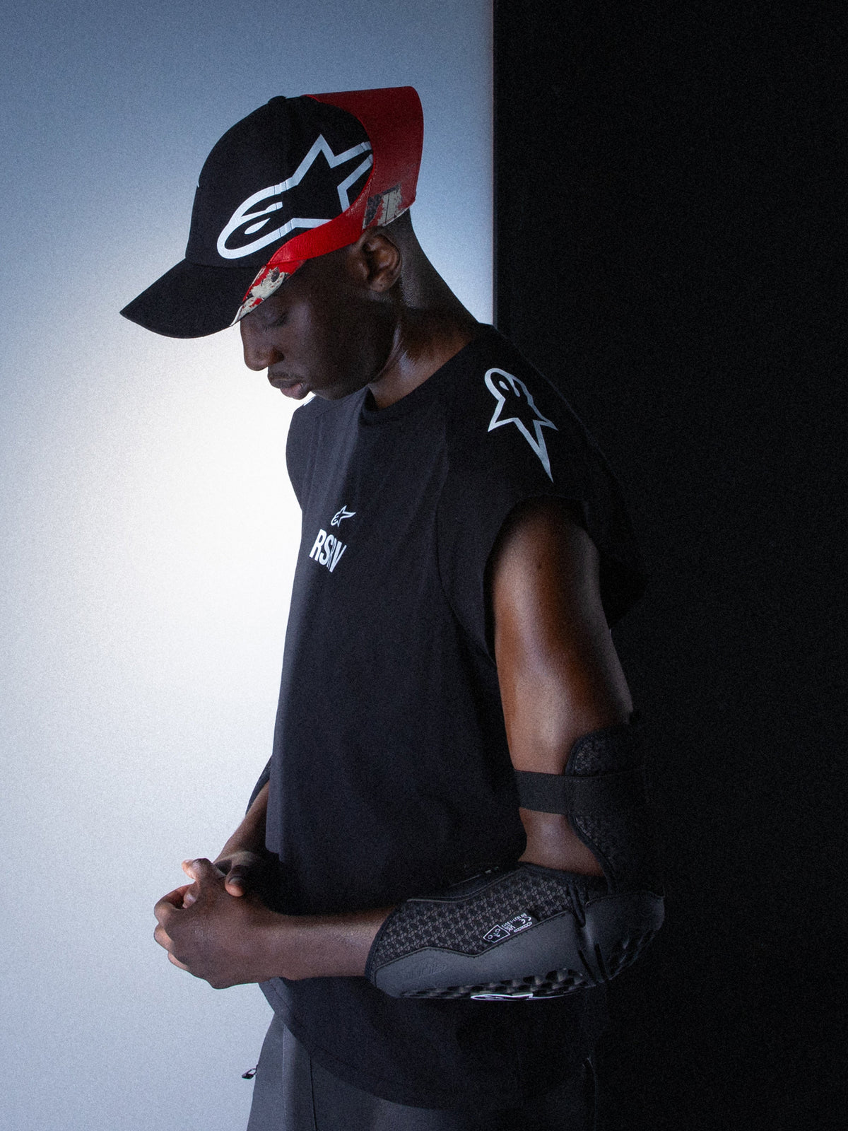 Alpinestars RSRV Tech Tank, camiseta sin mangas negra con logo blanco, persona usando gorra negra y roja, codera protectora negra con diseño texturizado, estilo urbano premium