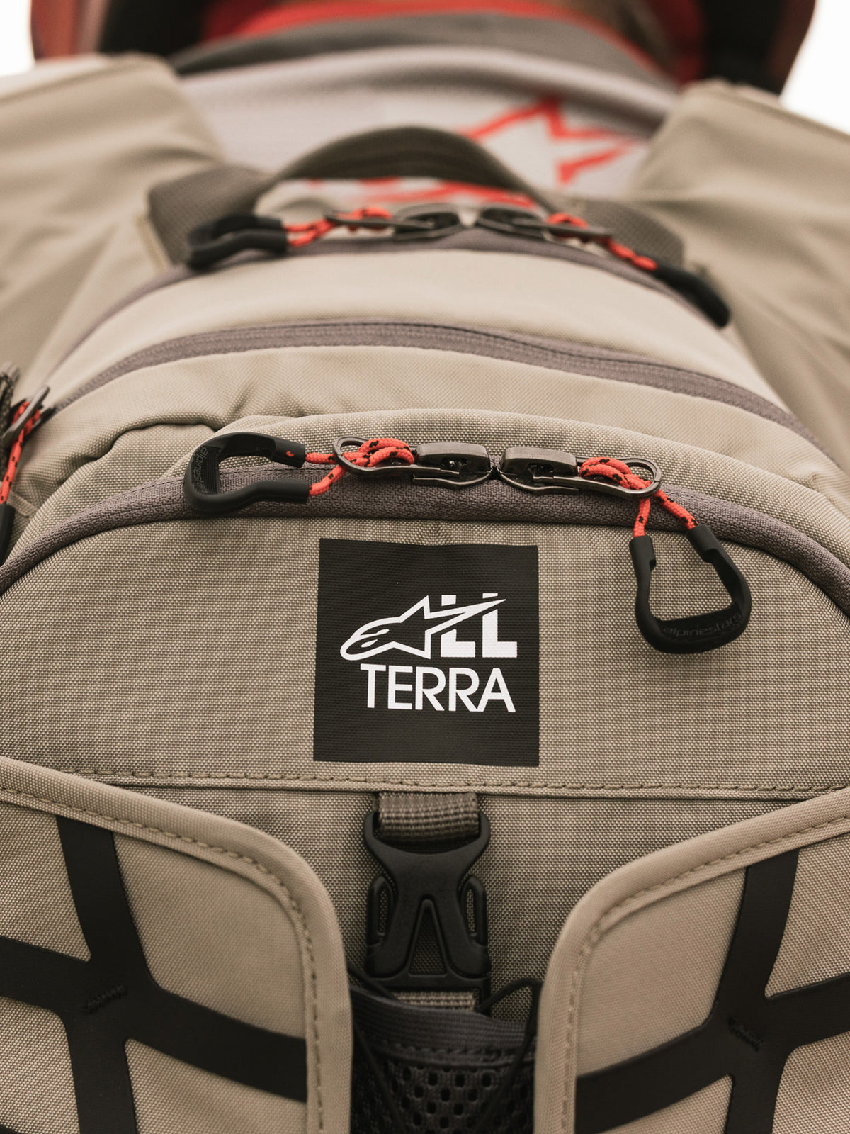 Mochila Alpinestars AT-15, Aluminum Walnut, primer plano del parche con el logotipo All Terra sobre tejido técnico color arena, con tiradores de cremallera de cordón naranja y detalles de construcción robusta
