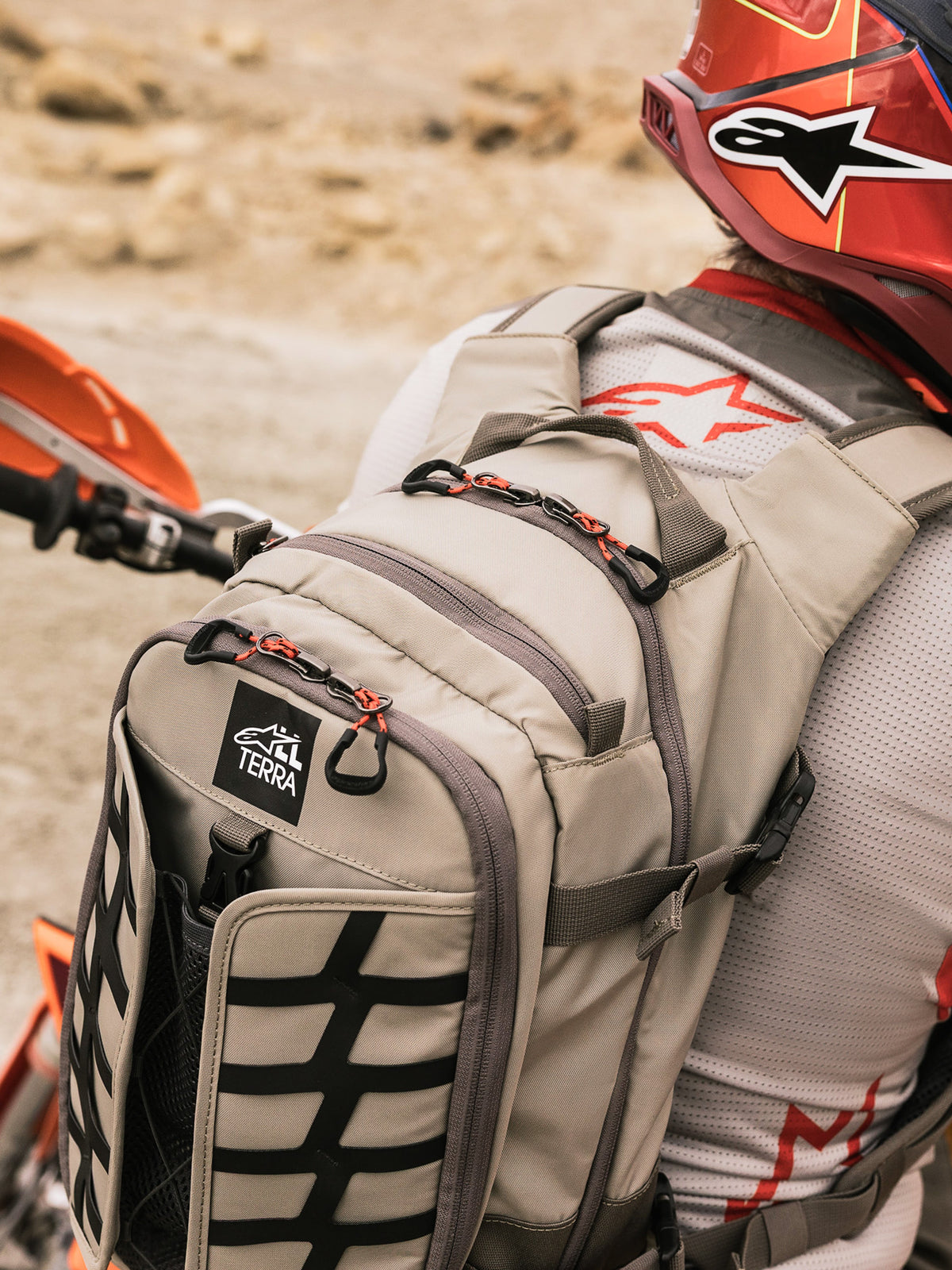 Mochila Alpinestars AT-15, mochila de moto para aventura, color nogal aluminio y negro, usada por un piloto en un entorno desértico, con tiradores de cremallera rojos, parche con el logo TERRA y correas externas negras, combinada con un casco rojo a juego y un jersey Alpinestars blanco