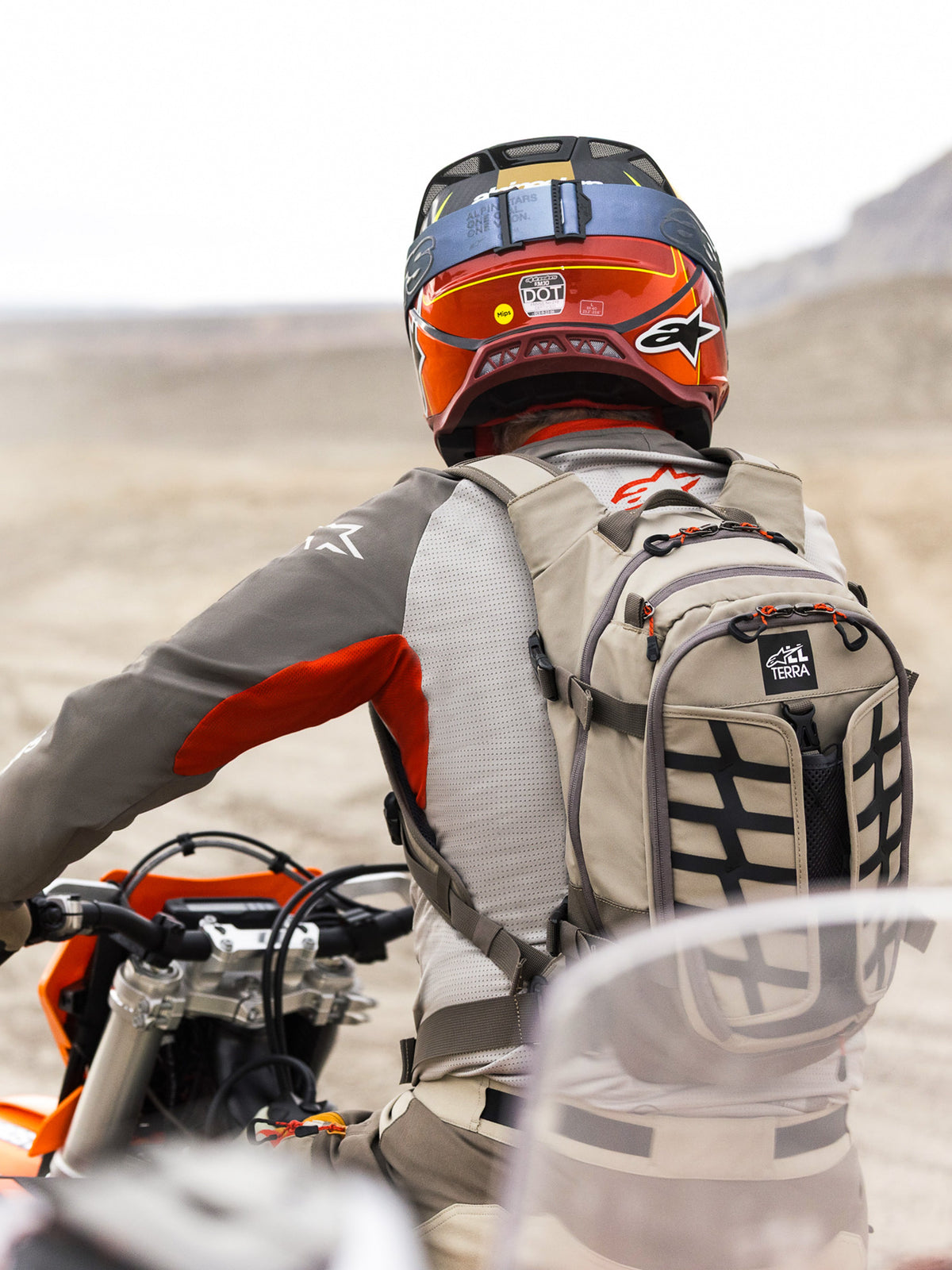 Mochila Alpinestars AT-15 en Aluminum Walnut, llevada por un motociclista de aventura en un paisaje desértico. Cuenta con una construcción textil beige resistente con correas negras, detalles naranjas en las cremalleras y el logotipo 'All Terra'. Combinada con un casco rojo y naranja y equipamiento gris para touring off-road.