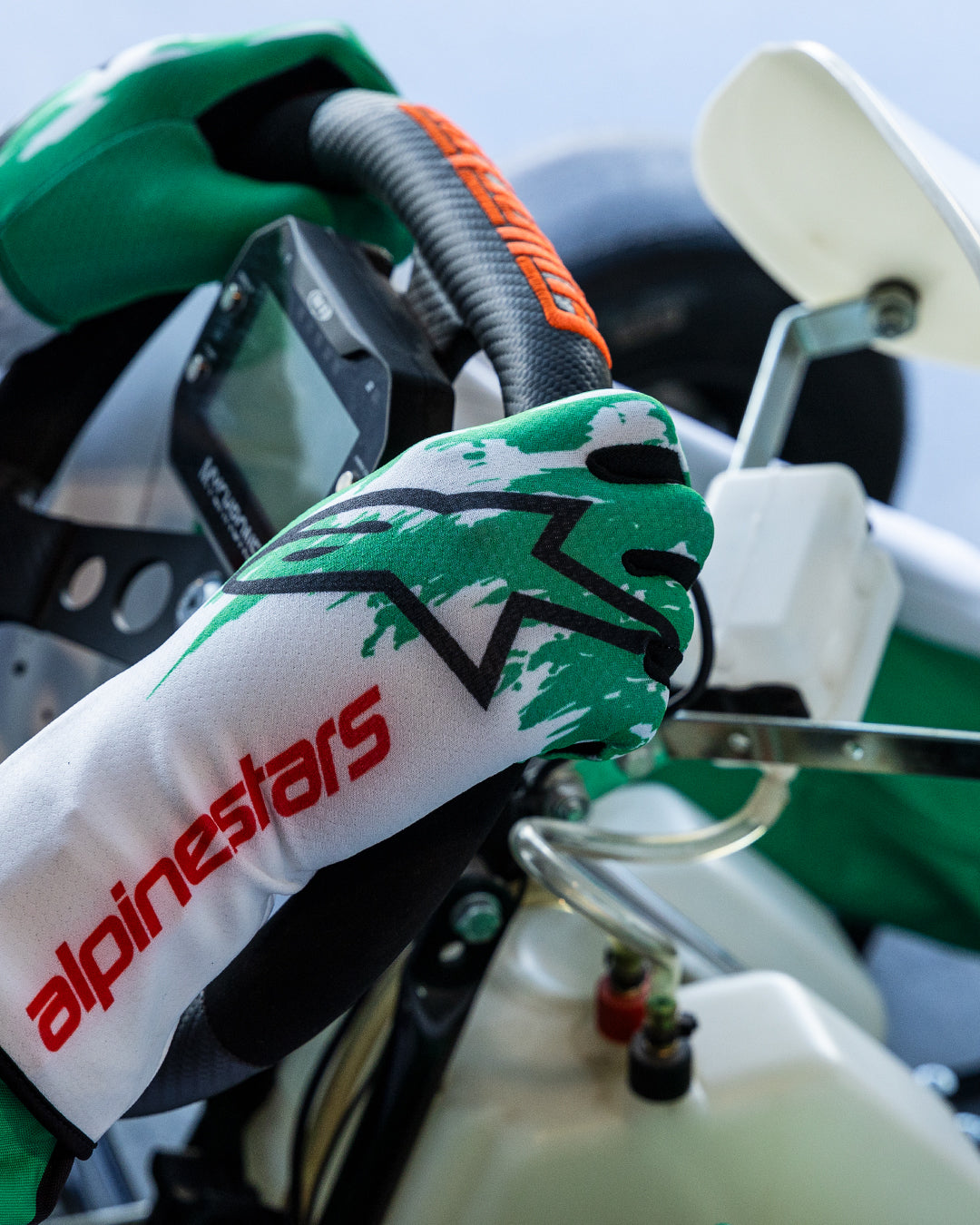 Guantes Alpinestars Tech-1 K V3 en blanco, verde y rojo, manos de un piloto sujetando un volante de karting, con un diseño de salpicaduras verdes sublimadas, logo de estrella negro y texto Alpinestars en rojo en el puño, equipamiento profesional para karting