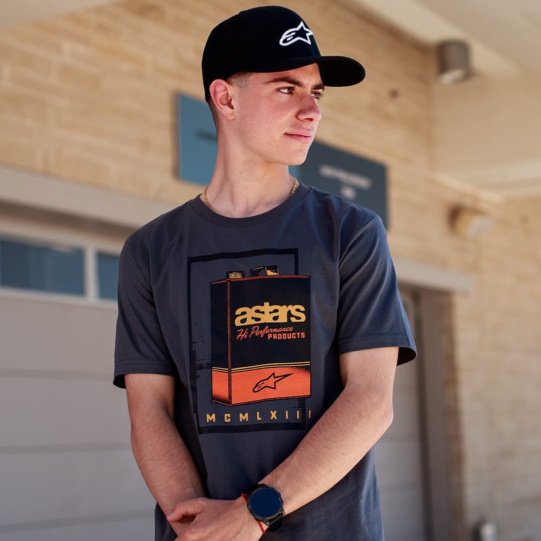 Alpinestars Galun CSF Tee, camiseta de manga corta, gris carbón, gráfico frontal de lata de gasolina vintage con marca 'astars' y números romanos 'MCMLXIII', usada por un hombre con gorra negra con logo, estilo de vida casual de motor