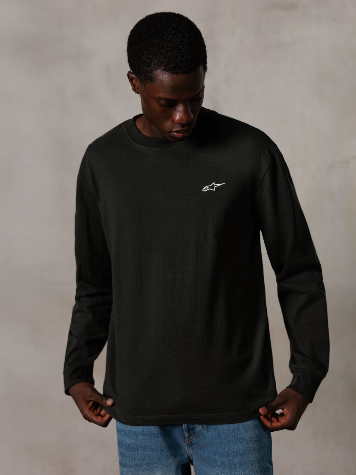 Alpinestars Essentials Ageless LS Tee, camiseta de manga larga, negro, con un pequeño logotipo blanco en el pecho, usada por un modelo con vaqueros azules, estilo urbano casual lifestyle