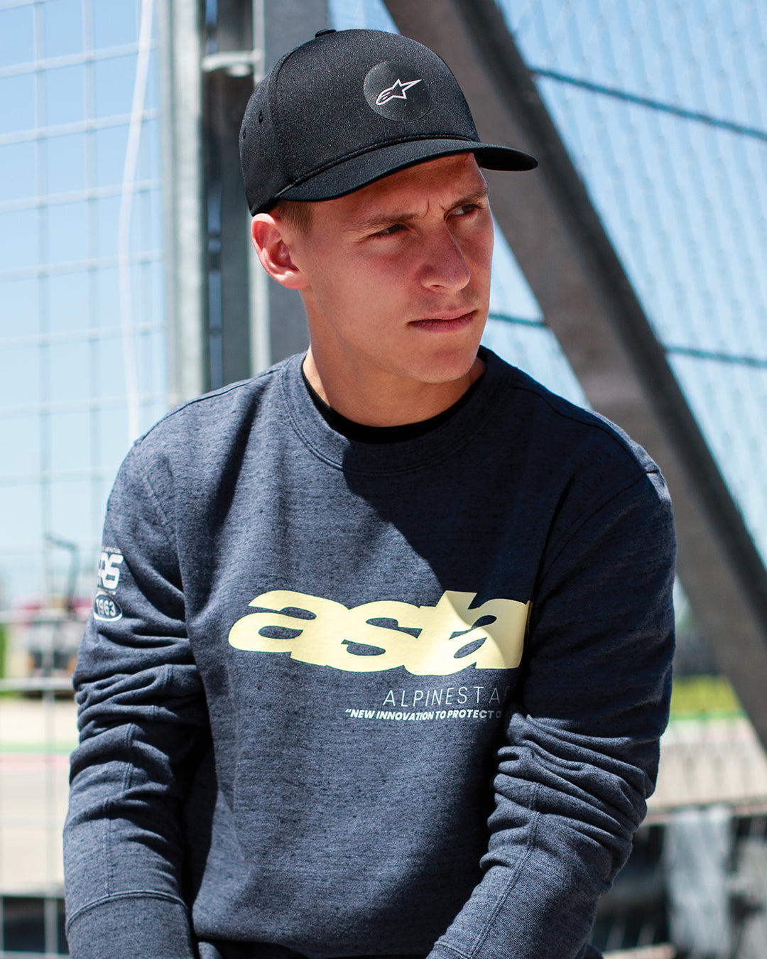 Fabio Quartararo con la gorra Alpinestars Apex en negro, gorra de copa estructurada con visera curva y logotipo de estrella circular, combinada con una sudadera gris oscuro jaspeado con marca en amarillo, estilo de vida casual inspirado en el motorsport