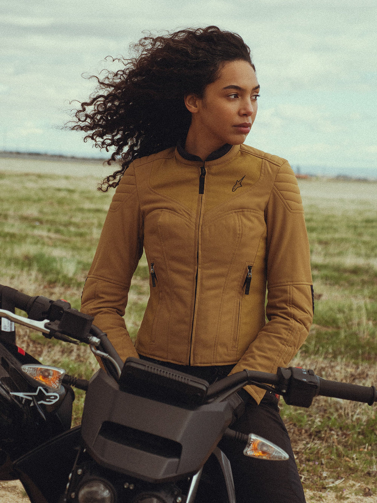 Chaqueta para mujer Alpinestars Isla WR en color Utility Brown Light Gold, lucida por una mujer sentada en una moto en un campo, con detalles acolchados en los hombros, silueta ajustada y estilo urbano contemporáneo.