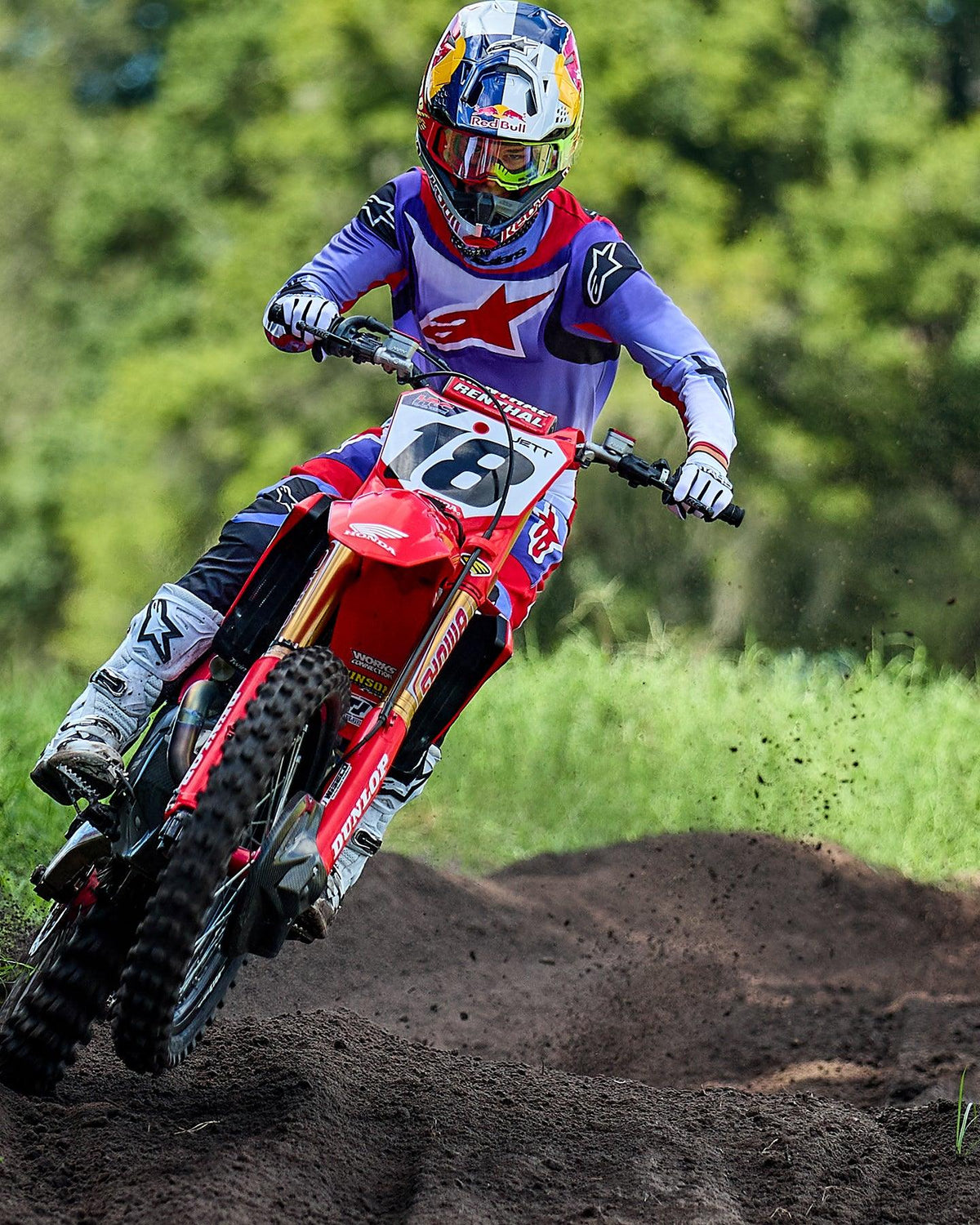 Piloto de motocross en una moto Honda roja, vistiendo el jersey Alpinestars 2025 Fluid Wurx en púrpura y rojo brillante, pantalones a juego, botas blancas y casco de Red Bull, recorriendo una curva en una pista de tierra.