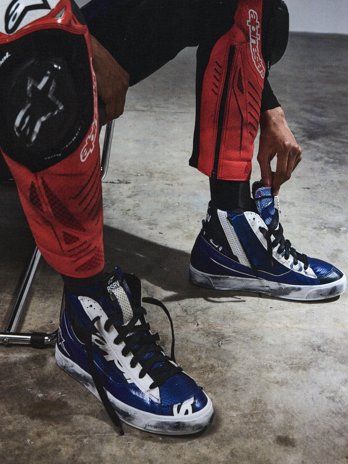 Alpinestars RSRV Legacy Shoe, zapatillas altas azules y blancas con cordones negros, persona ajustándose las zapatillas mientras usa pantalones protectores rojos y negros, superficie de concreto gris