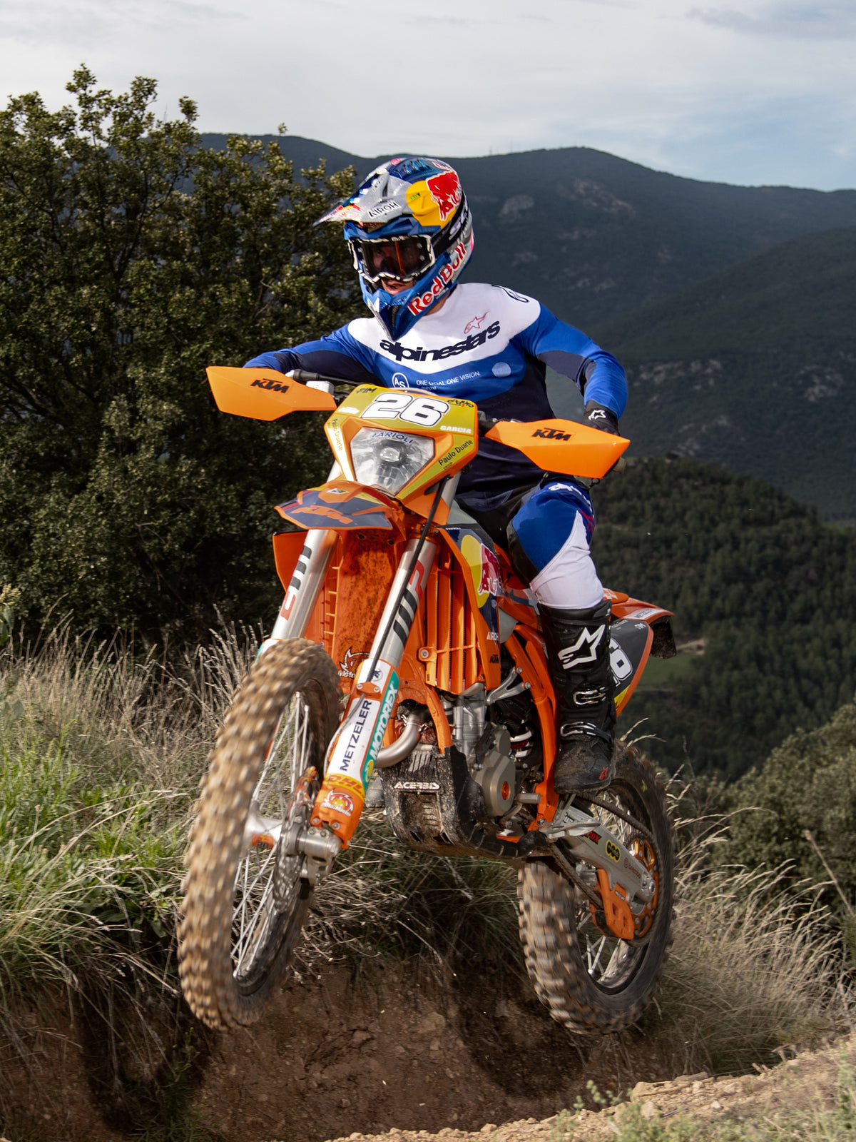 Piloto de enduro en una moto de cross naranja, con Jersey Alpinestars Pro-Dura en azul marino, azul brillante y blanco, pantalones de montar negros y blancos, y botas Alpinestars negras, recorriendo un sendero escarpado con montañas y árboles al fondo.