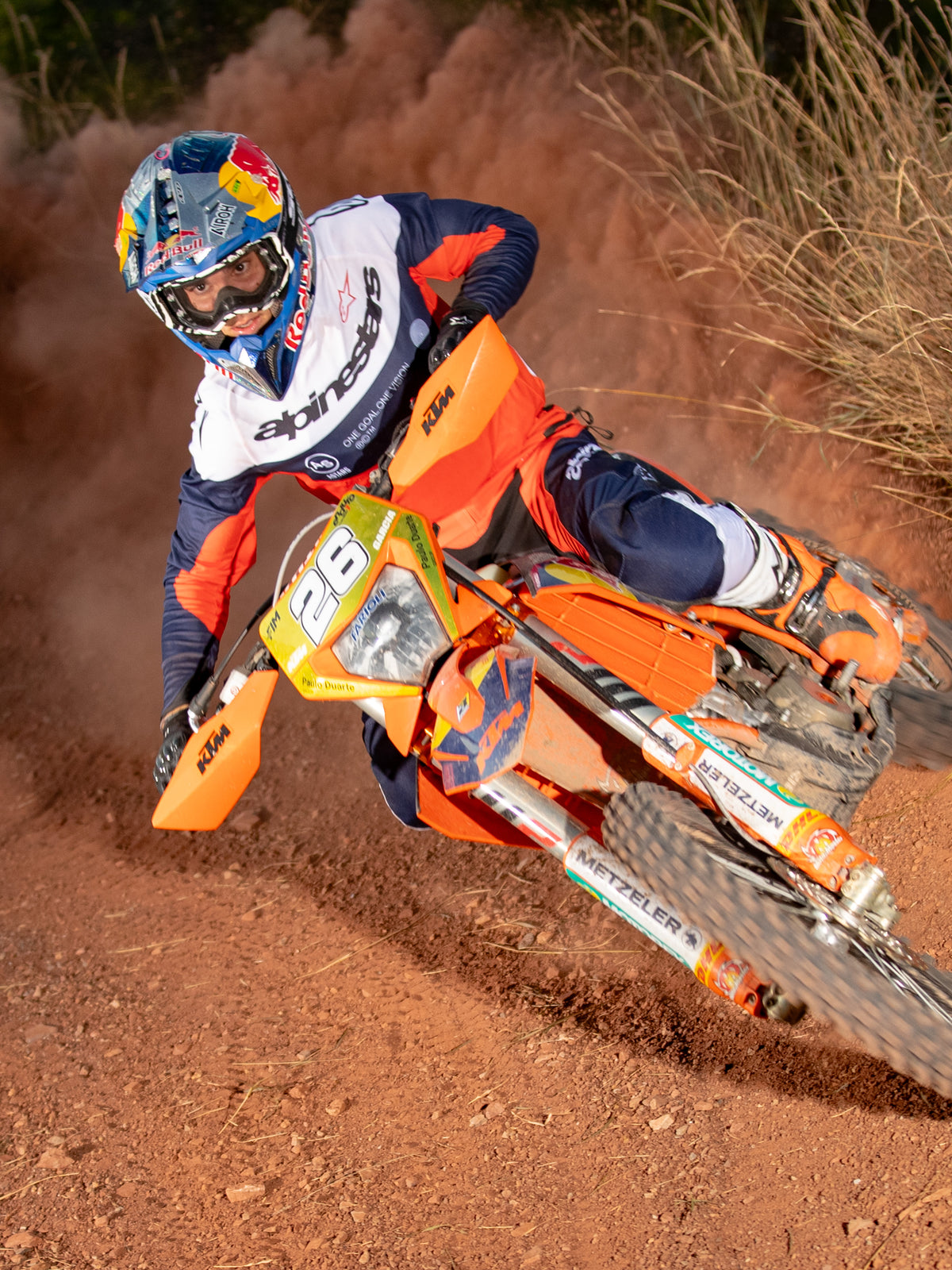 Piloto de motocross inclinándose en una curva en una motocicleta naranja, vistiendo jersey y pantalones Alpinestars Pro-Dura en azul navy, naranja hot y blanco, equipo de carreras off-road coordinado, casco con marca Red Bull, botas blancas, toma de acción en una pista de tierra polvorienta.
