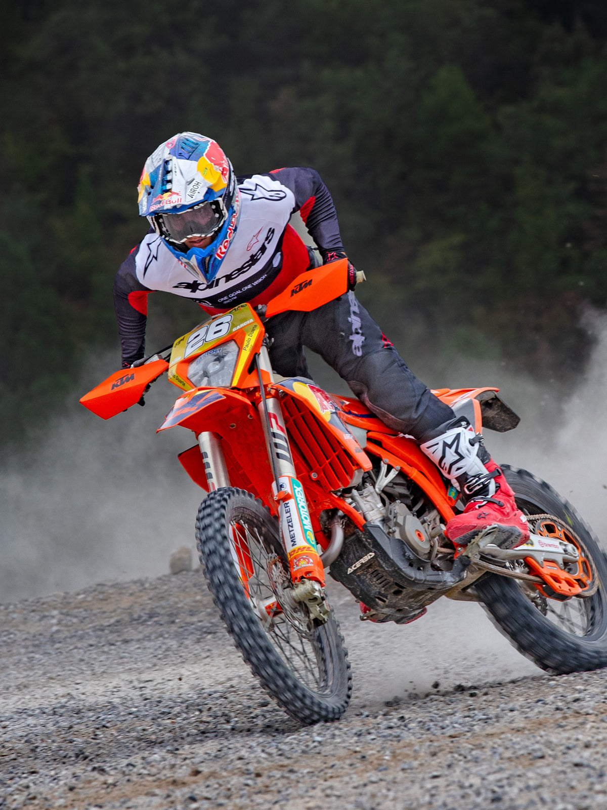 Piloto de motocross en acción vistiendo el Jersey Alpinestars Pro-Dura en negro, rojo brillante y blanco, conduciendo una moto de cross naranja en un sendero todoterreno polvoriento, combinado con pantalones negros y botas rojas y blancas