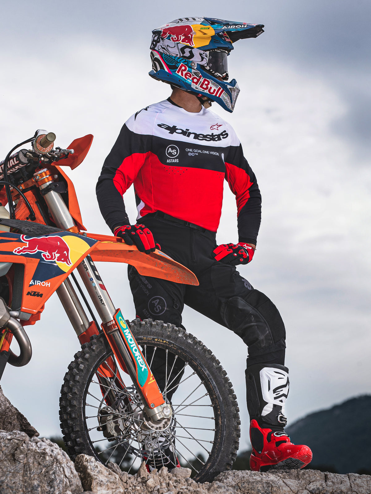 Piloto de motocross de pie junto a una motocicleta todoterreno naranja en terreno rocoso, vistiendo equipamiento Alpinestars Pro-Dura con jersey rojo, blanco y negro y pantalones negros, complementado con guantes rojos y negros, botas blancas y rojas, y casco de Red Bull, conjunto profesional de enduro