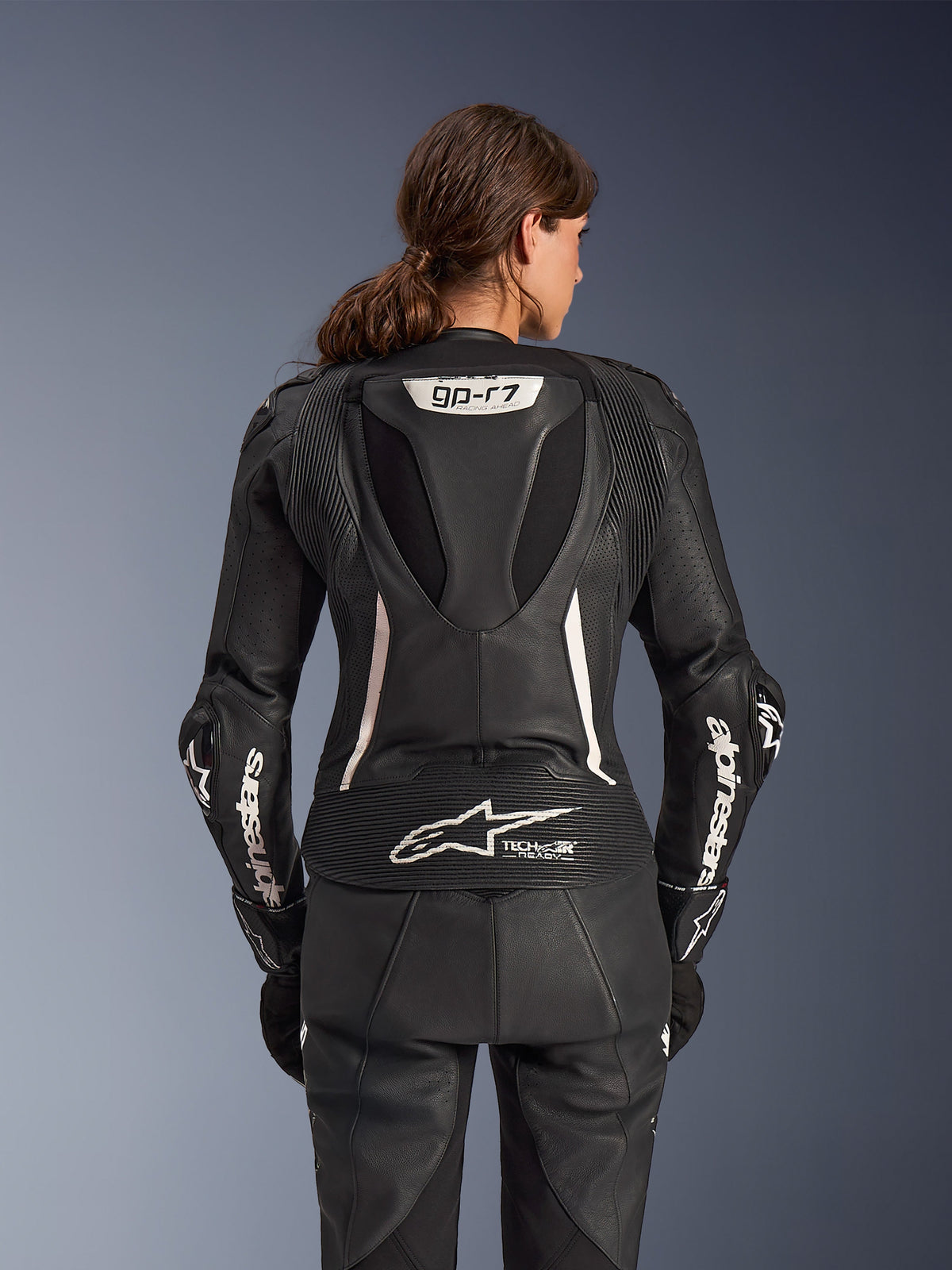 Traje de cuero de 2 piezas Alpinestars Stella GP-R7, traje de moto para mujer, negro con detalles blancos, vista posterior que muestra la joroba aerodinámica Supertech R10 con el logo GP-R7, marca Tech-Air Ready en la parte inferior de la espalda, letras Alpinestars blancas en las mangas y paneles elásticos de acordeón para un alto rendimiento en competición
