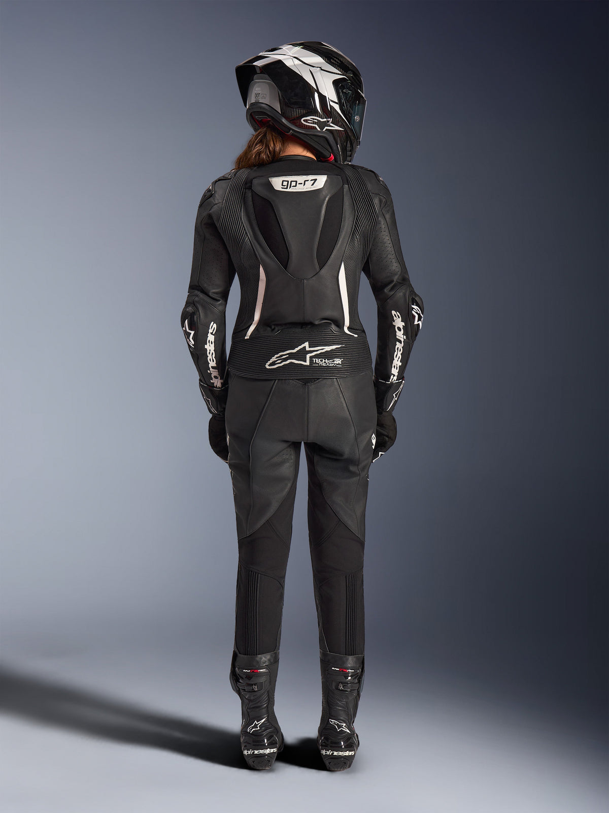 Traje de piel de 2 piezas Alpinestars Stella GP-R7 para mujer, negro con detalles blancos, vista trasera que muestra la joroba aerodinámica Supertech R10 y el logotipo Tech-Air Ready, usado con casco integral a juego, guantes de carreras y botas de piel para conducción en pista y deportiva