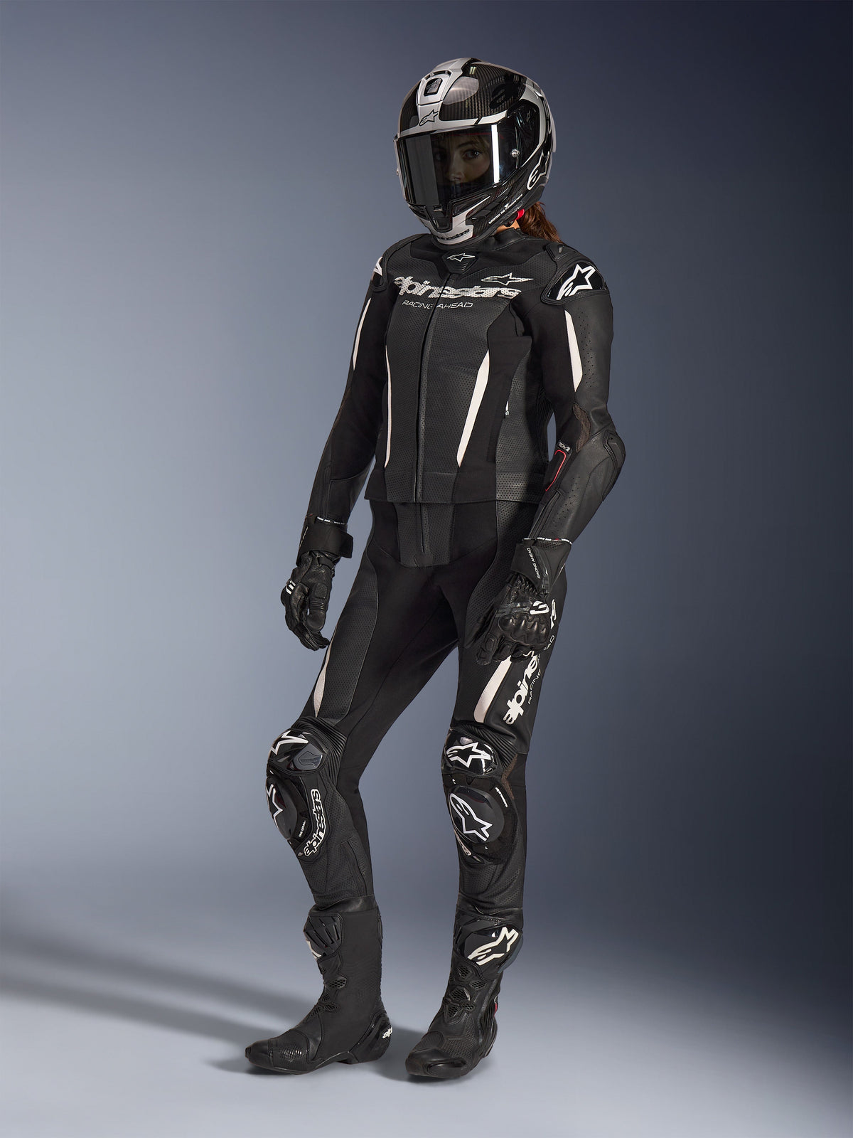 Traje de cuero de 2 piezas Alpinestars Stella GP-R7, traje de motocicleta para mujer, negro con detalles en blanco, usado por una piloto con casco de carreras integral, guantes negros y botas, con paneles de cuero perforado y logotipos de Alpinestars en blanco, equipamiento profesional de carreras y alto rendimiento deportivo