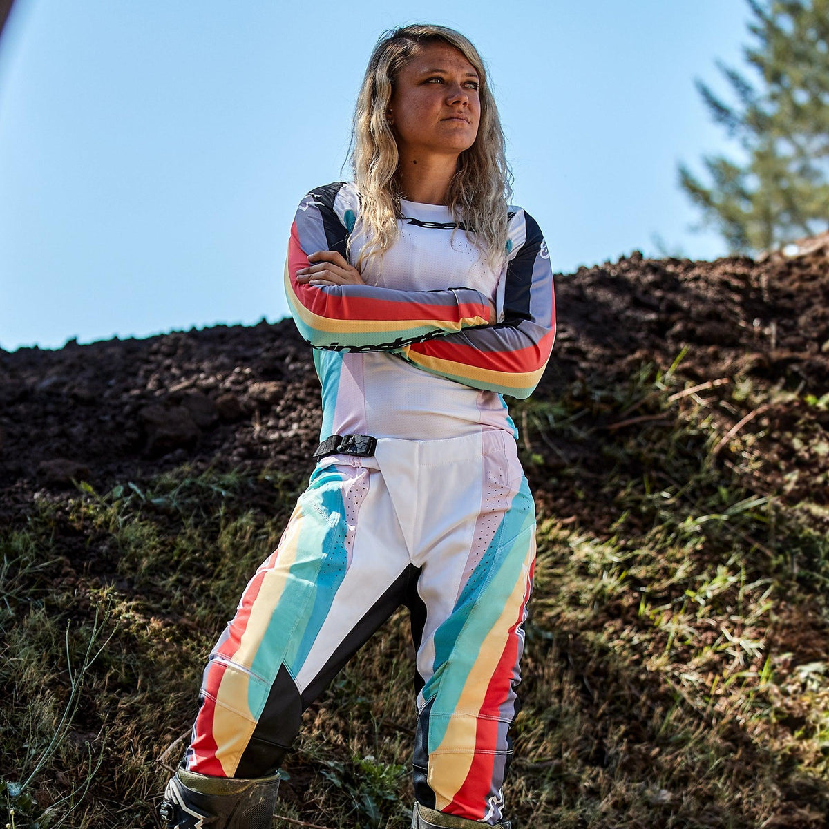 Alpinestars Stella Techstar Pants 2024 para mujer, pantalones de motocross con jersey a juego, diseño Black Multicolor con base blanca y rayas vibrantes en azul, amarillo y rojo, piloto femenina de pie con los brazos cruzados en una pista de tierra, equipamiento profesional de carreras off-road