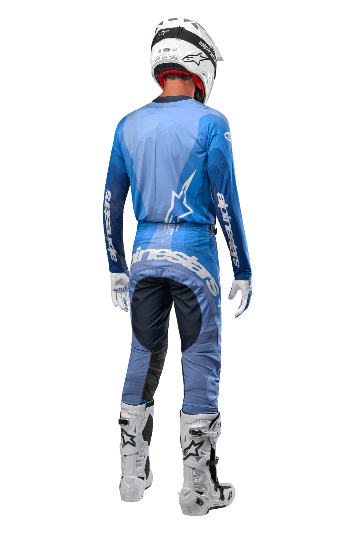 Piloto de motocross visto desde atrás con los pantalones Alpinestars Techstar Pneuma 2024 en azul marino y azul claro, combinados con un jersey azul a juego, casco blanco, guantes blancos y botas blancas, con la marca Alpinestars destacada en el asiento y las mangas.