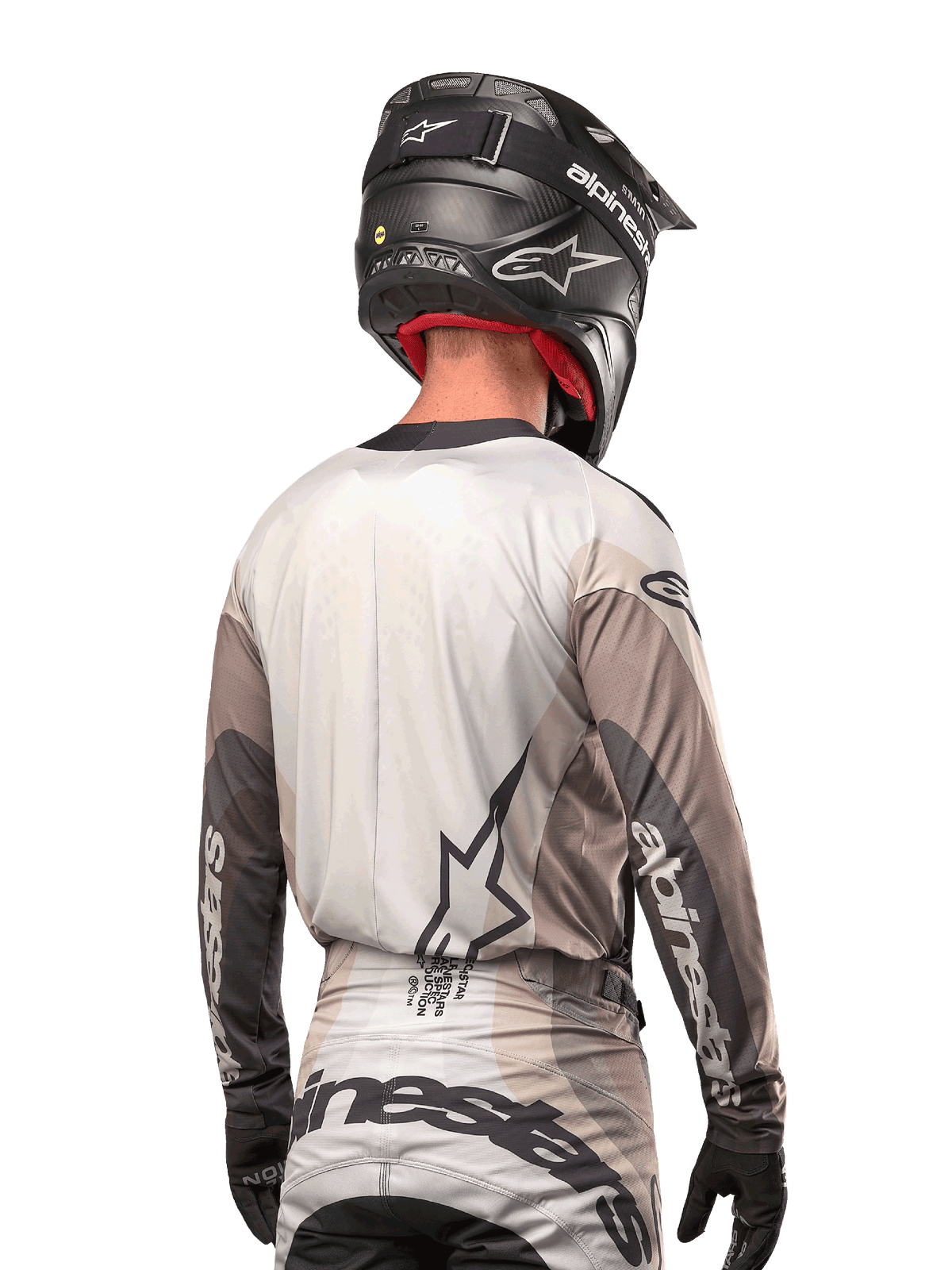 Piloto de motocross visto desde atrás con el Jersey Alpinestars Techstar Pneuma 2024 en gris arena y gris hierro, combinado con un casco negro con acabado de carbono, con un gran logo de estrella en la espalda y la marca Alpinestars en las mangas.