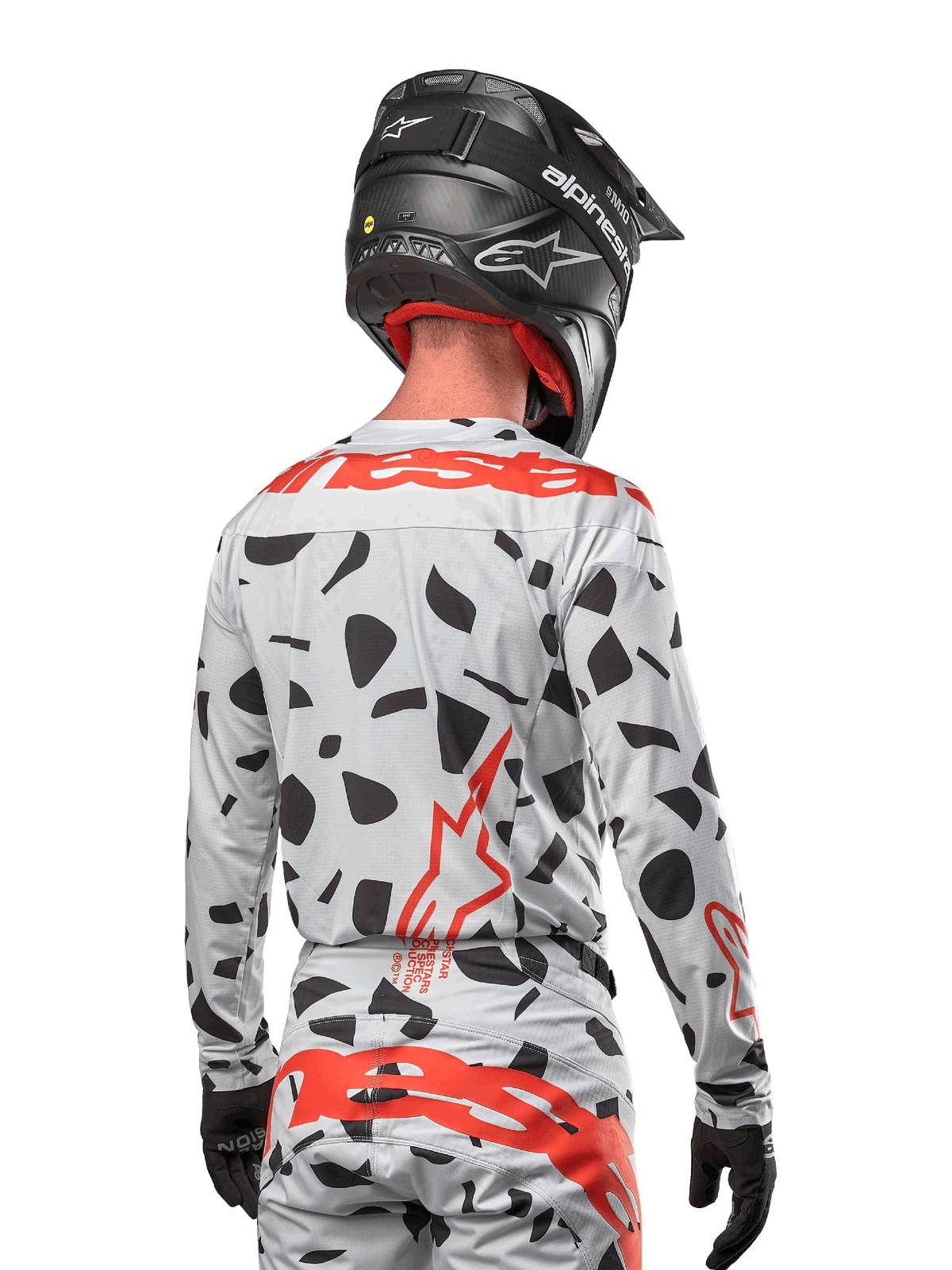 Piloto de motocross desde atrás vistiendo el jersey Alpinestars 2024 Techstar Rantera en Haze Gray Camo, con una base gris claro con manchas gráficas negras y logotipos rojos destacados de Alpinestars en la espalda y hombros, combinado con pantalones a juego y un casco negro.