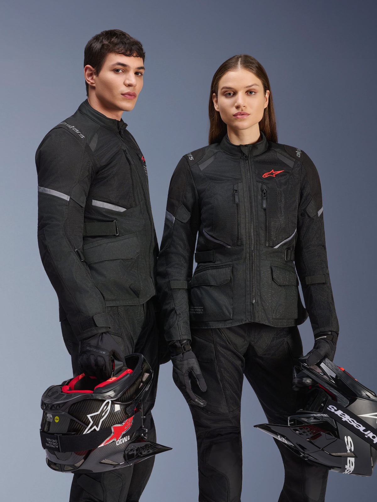 Male and female models wearing Alpinestars Stella Andes Air Drystar® Jackets in Black, adventure motorcycle touring gear featuring red star logos, mesh ventilation panels, and technical textile construction, models holding adventure helmets in a studio shot