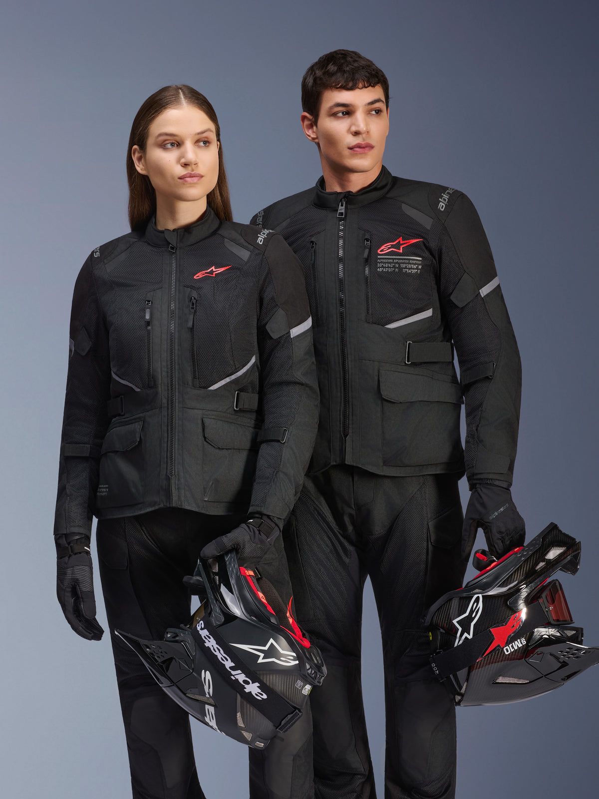 Man and woman wearing Alpinestars Andes Air Drystar® Jackets, Black adventure touring gear with mesh panels and reflective details, matching black pants and gloves, holding motocross helmets, studio shot of professional motorcycle apparel