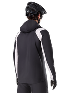 Alpinestars A-Dura Rain Jacket, chaqueta impermeable para ciclismo de montaña, negra con paneles blancos en las mangas, vista trasera de un ciclista con casco de MTB y guantes negros, construcción técnica impermeable de 2.5 capas, ropa de ciclismo de montaña