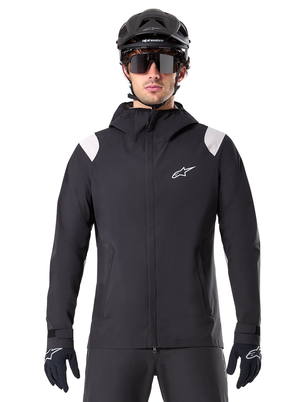 Alpinestars A-Dura Rain Jacket, chaqueta de lluvia para MTB, negra con paneles blancos en los hombros, usada con casco de MTB negro, gafas de sol y guantes, diseño impermeable con capucha y cremallera frontal, equipamiento deportivo para ciclismo