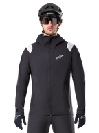 Alpinestars A-Dura Rain Jacket, chaqueta de lluvia para MTB, negra con paneles blancos en los hombros, usada con casco de MTB negro, gafas de sol y guantes, diseño impermeable con capucha y cremallera frontal, equipamiento deportivo para ciclismo