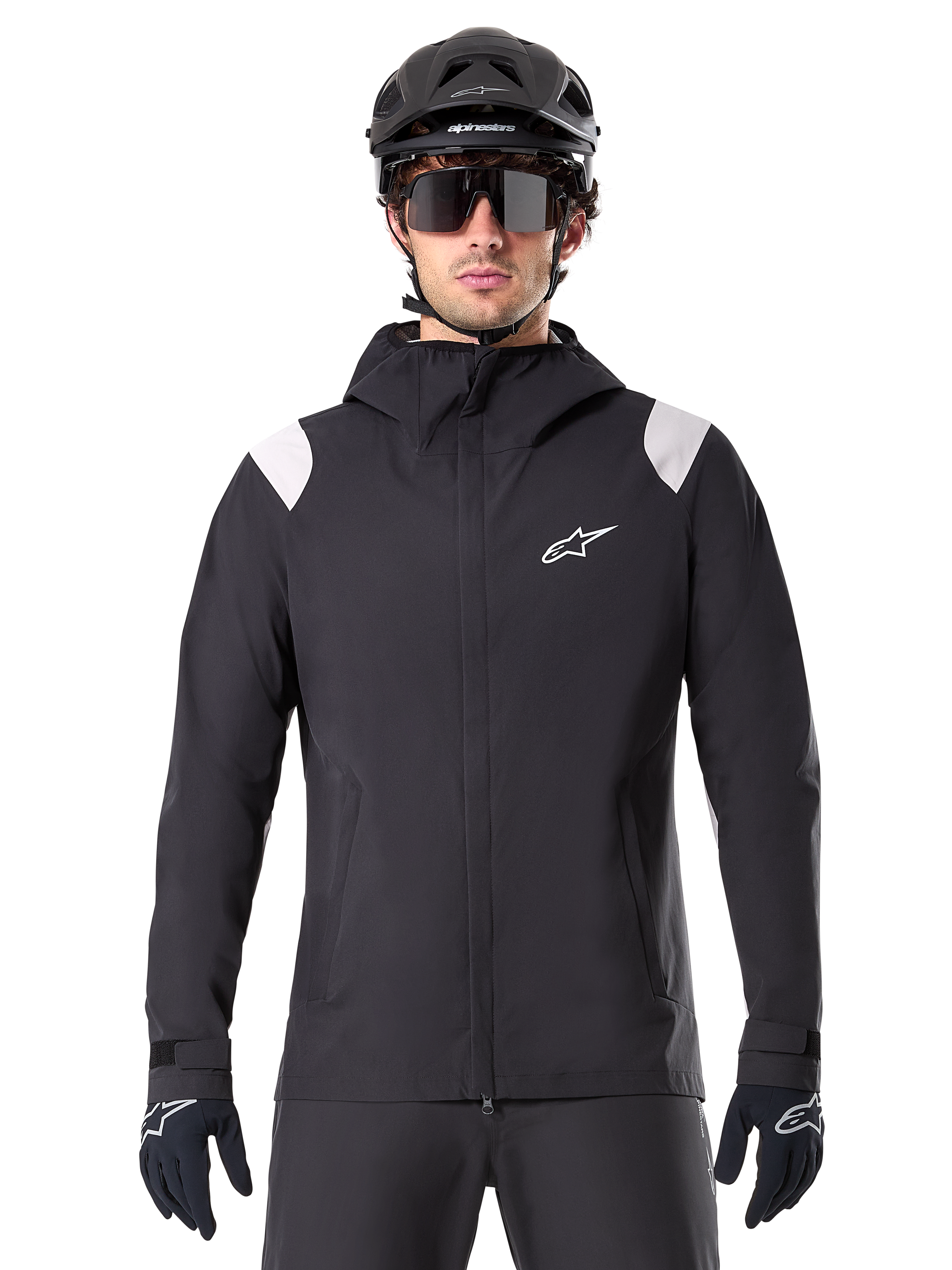 Alpinestars A-Dura Rain Jacket, chaqueta de lluvia para MTB, negra con paneles blancos en los hombros, usada con casco de MTB negro, gafas de sol y guantes, diseño impermeable con capucha y cremallera frontal, equipamiento deportivo para ciclismo