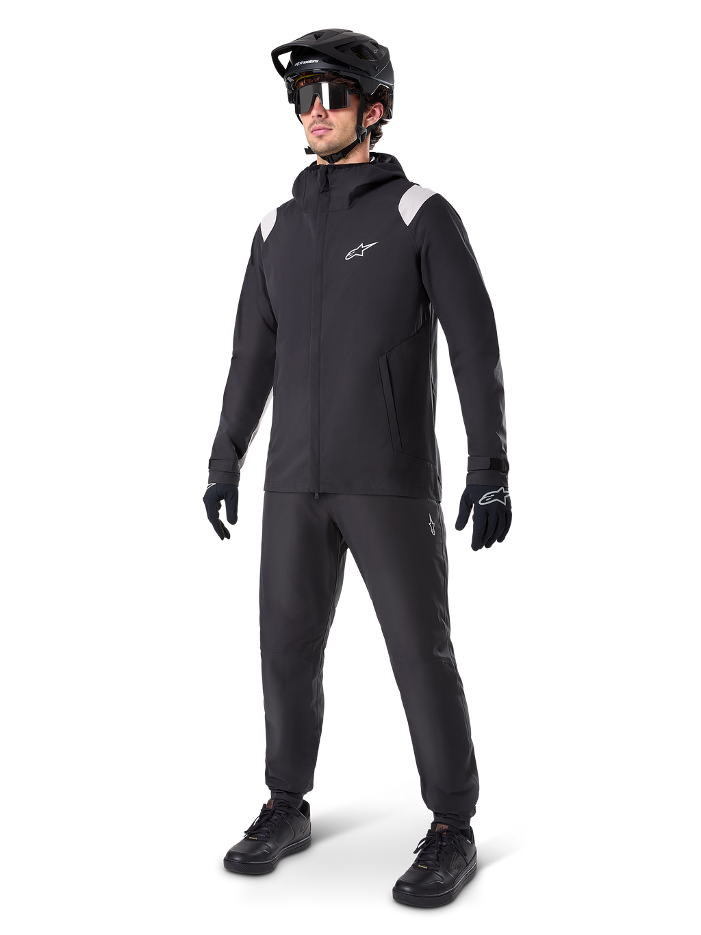 Alpinestars Chaqueta de lluvia A-Dura, chaqueta impermeable negra para ciclismo de montaña, con detalles blancos en los hombros, lucida por un ciclista con casco de MTB negro a juego, gafas de sol, guantes y pantalones, equipamiento técnico de ciclismo para todo tipo de clima