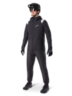 Alpinestars Chaqueta de lluvia A-Dura, chaqueta impermeable negra para ciclismo de montaña, con detalles blancos en los hombros, lucida por un ciclista con casco de MTB negro a juego, gafas de sol, guantes y pantalones, equipamiento técnico de ciclismo para todo tipo de clima