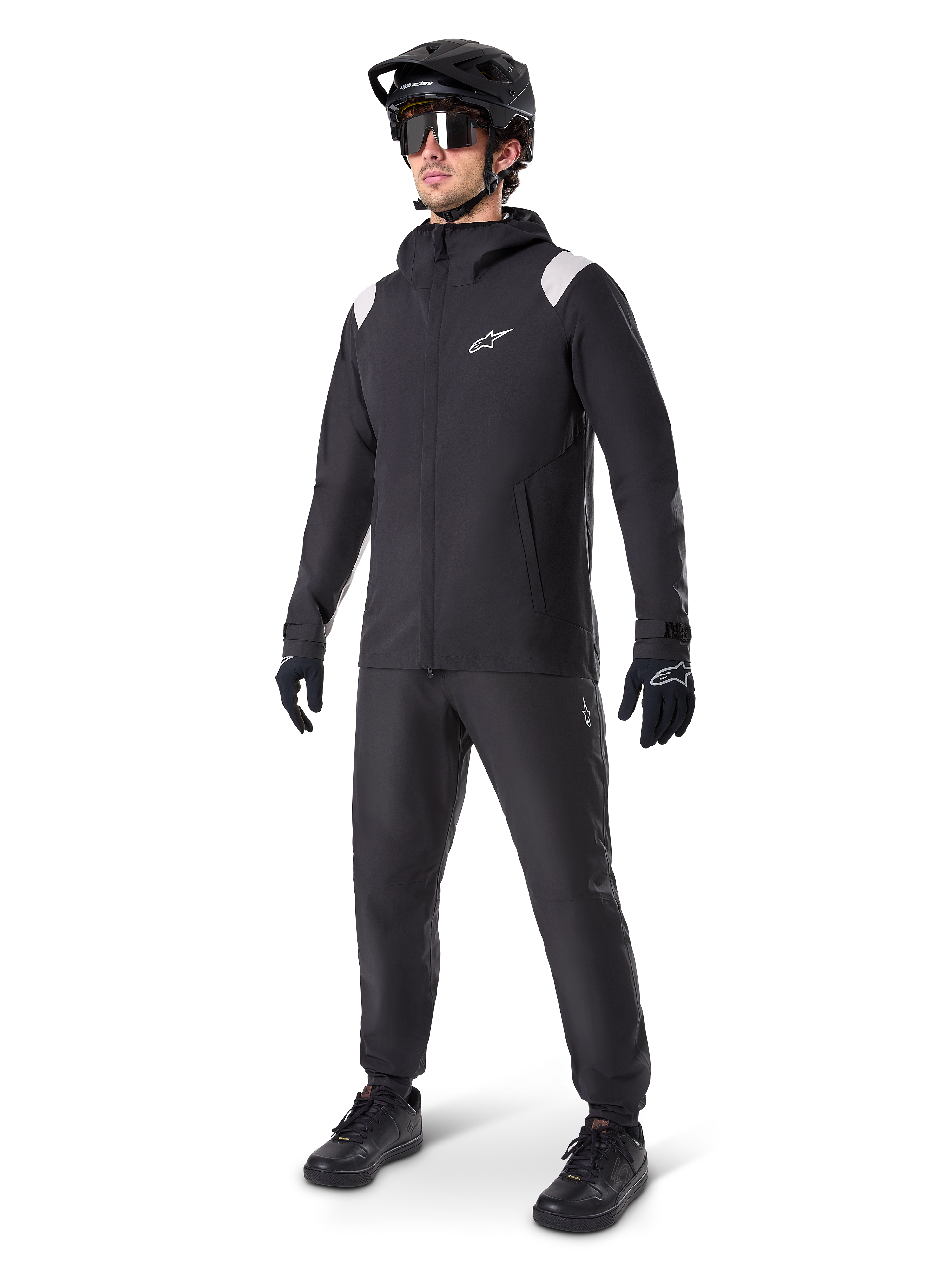 Alpinestars Chaqueta de lluvia A-Dura, chaqueta impermeable negra para ciclismo de montaña, con detalles blancos en los hombros, lucida por un ciclista con casco de MTB negro a juego, gafas de sol, guantes y pantalones, equipamiento técnico de ciclismo para todo tipo de clima