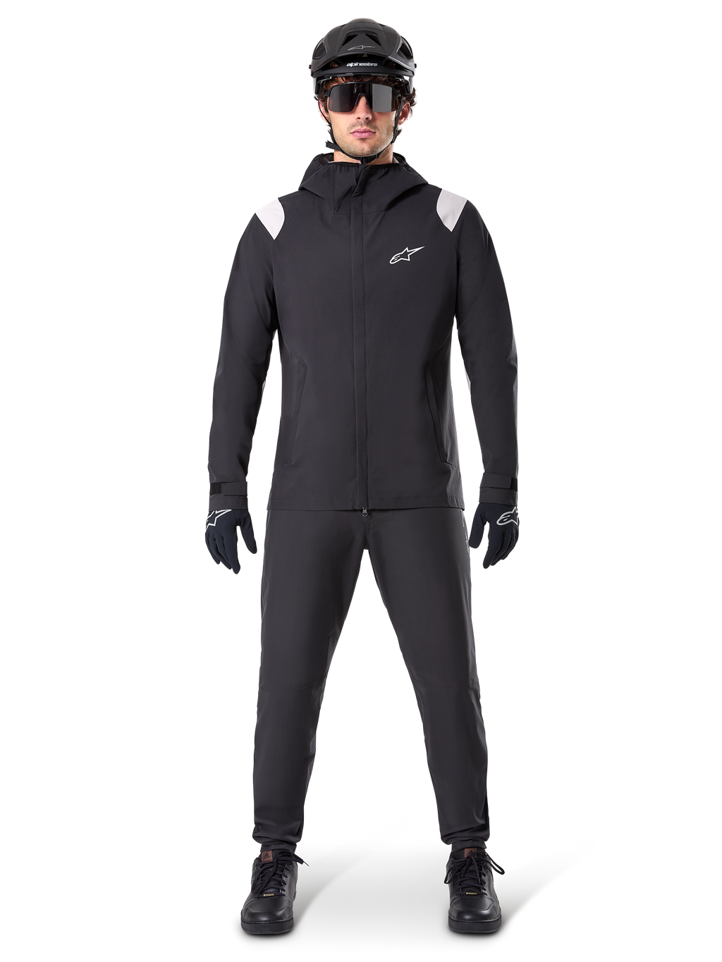 Chaqueta impermeable Alpinestars A-Dura, chaqueta de lluvia para mountain bike, negra con detalles blancos en los hombros y logotipo blanco en el pecho, usada con pantalones negros, casco negro, gafas de sol y guantes, equipamiento de ciclismo de montaña coordinado para clima húmedo.