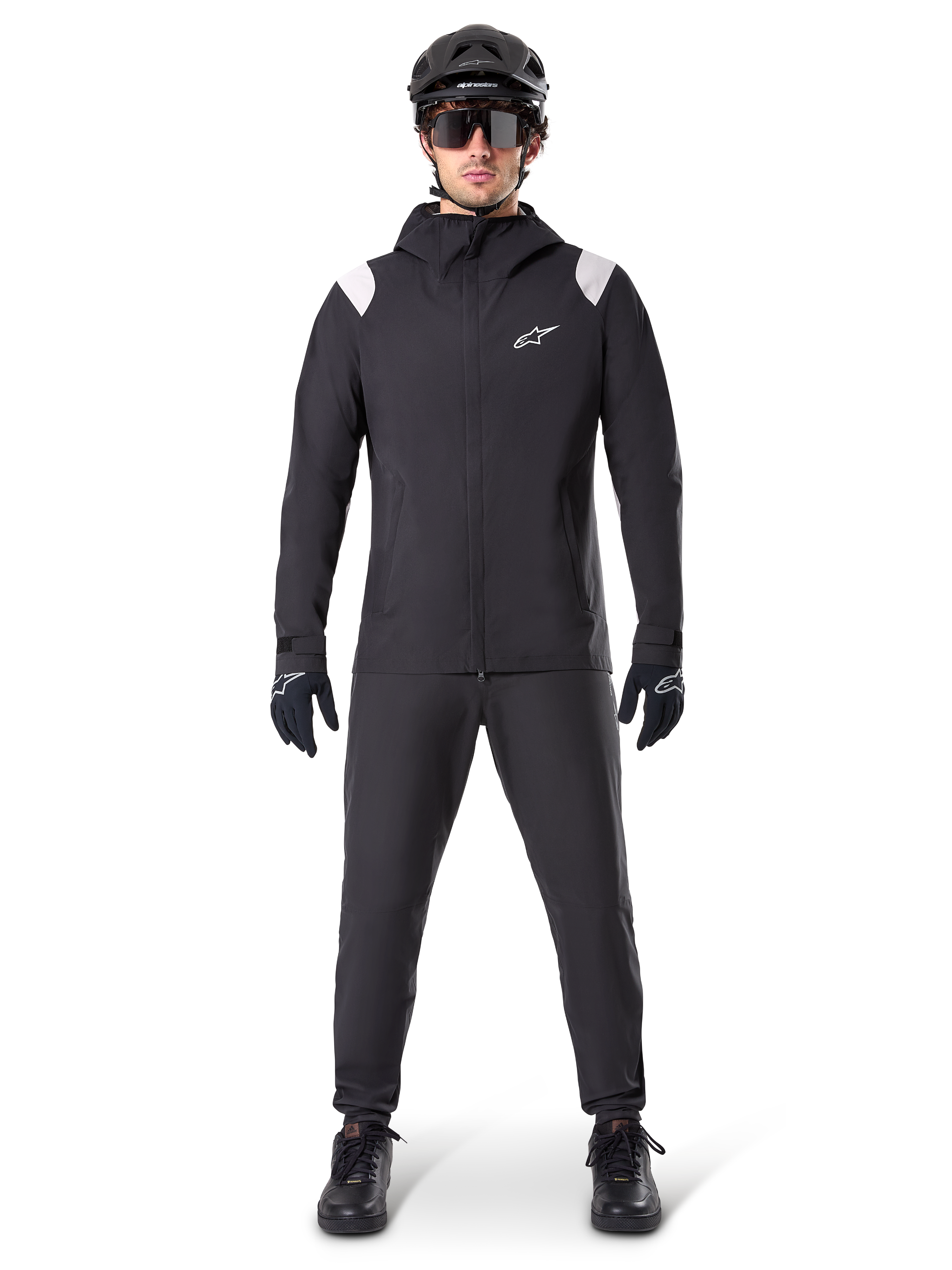 Chaqueta impermeable Alpinestars A-Dura, chaqueta de lluvia para mountain bike, negra con detalles blancos en los hombros y logotipo blanco en el pecho, usada con pantalones negros, casco negro, gafas de sol y guantes, equipamiento de ciclismo de montaña coordinado para clima húmedo.
