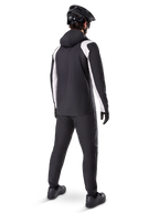 Alpinestars A-Dura Rain Jacket, vista trasera de un ciclista masculino con la chaqueta de MTB impermeable en negro y blanco, pantalones de ciclismo negros a juego, casco negro y guantes, equipamiento técnico para ciclismo de montaña en condiciones húmedas