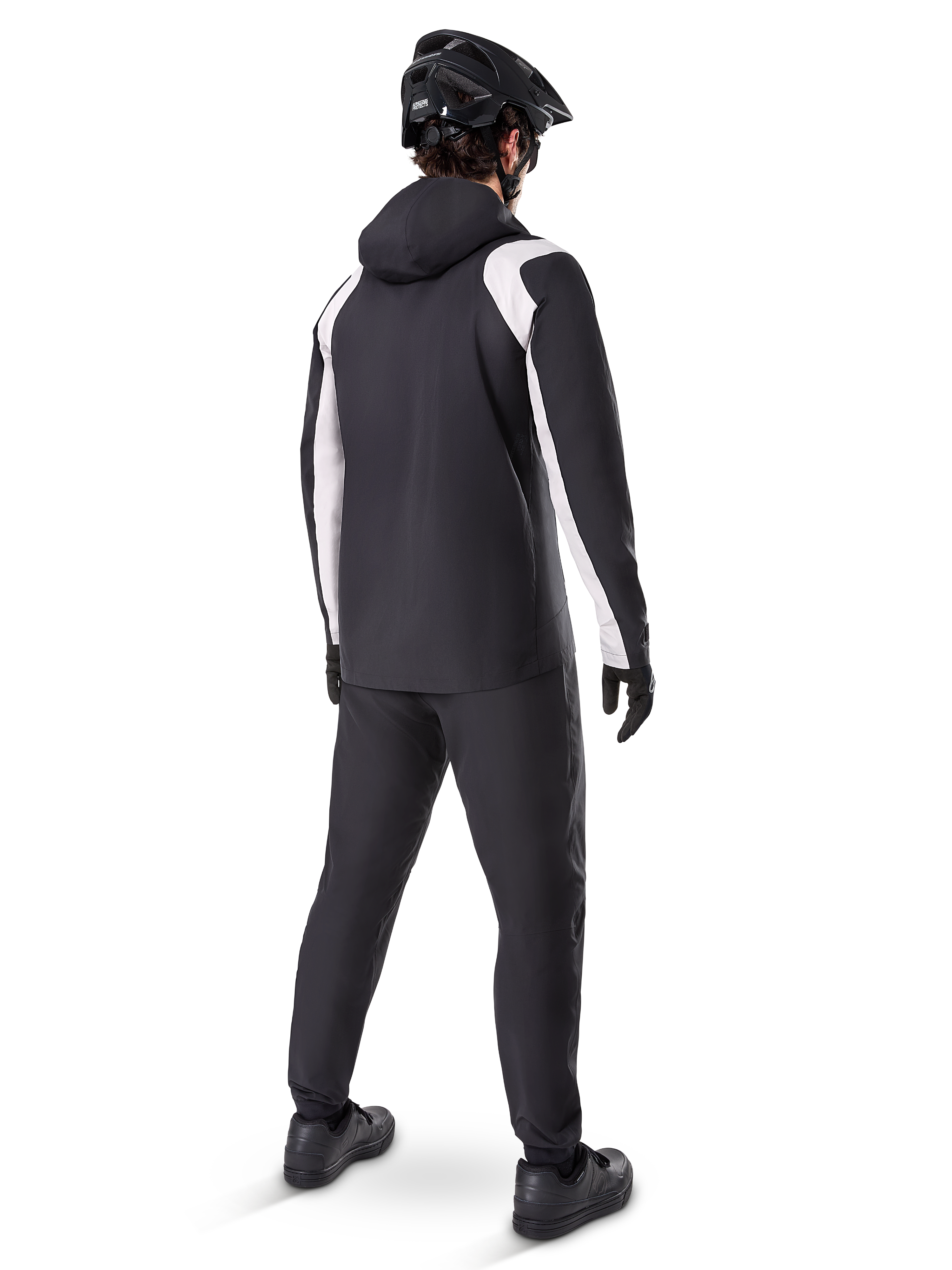 Alpinestars A-Dura Rain Jacket, vista trasera de un ciclista masculino con la chaqueta de MTB impermeable en negro y blanco, pantalones de ciclismo negros a juego, casco negro y guantes, equipamiento técnico para ciclismo de montaña en condiciones húmedas