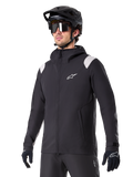 Alpinestars A-Dura Rain Jacket, chaqueta de lluvia para mountain bike, negra con detalles en los hombros en gris claro, usada por un ciclista con casco de MTB negro y gafas de sol, logo blanco en el pecho, construcción impermeable, coordinada con pantalones y guantes negros para ciclismo de montaña