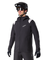 Alpinestars A-Dura Rain Jacket, chaqueta de lluvia para mountain bike, negra con detalles en los hombros en gris claro, usada por un ciclista con casco de MTB negro y gafas de sol, logo blanco en el pecho, construcción impermeable, coordinada con pantalones y guantes negros para ciclismo de montaña