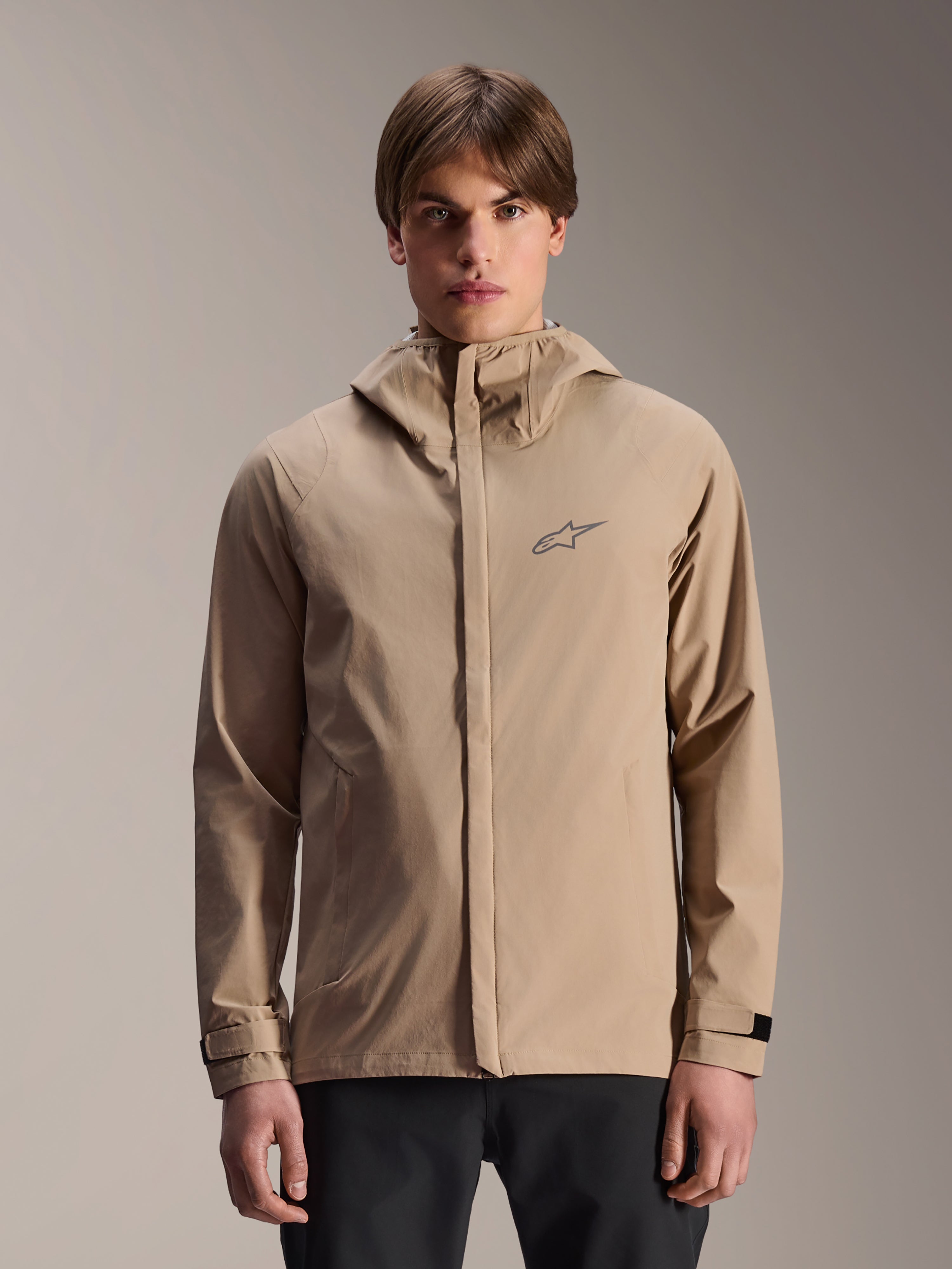 Alpinestars A-Dura Rain Jacket, chaqueta de lluvia con capucha para mountain bike en color marrón claro, usada por un modelo, con un pequeño logotipo en el pecho, correas de puño ajustables y cremallera frontal oculta, equipo de ciclismo impermeable
