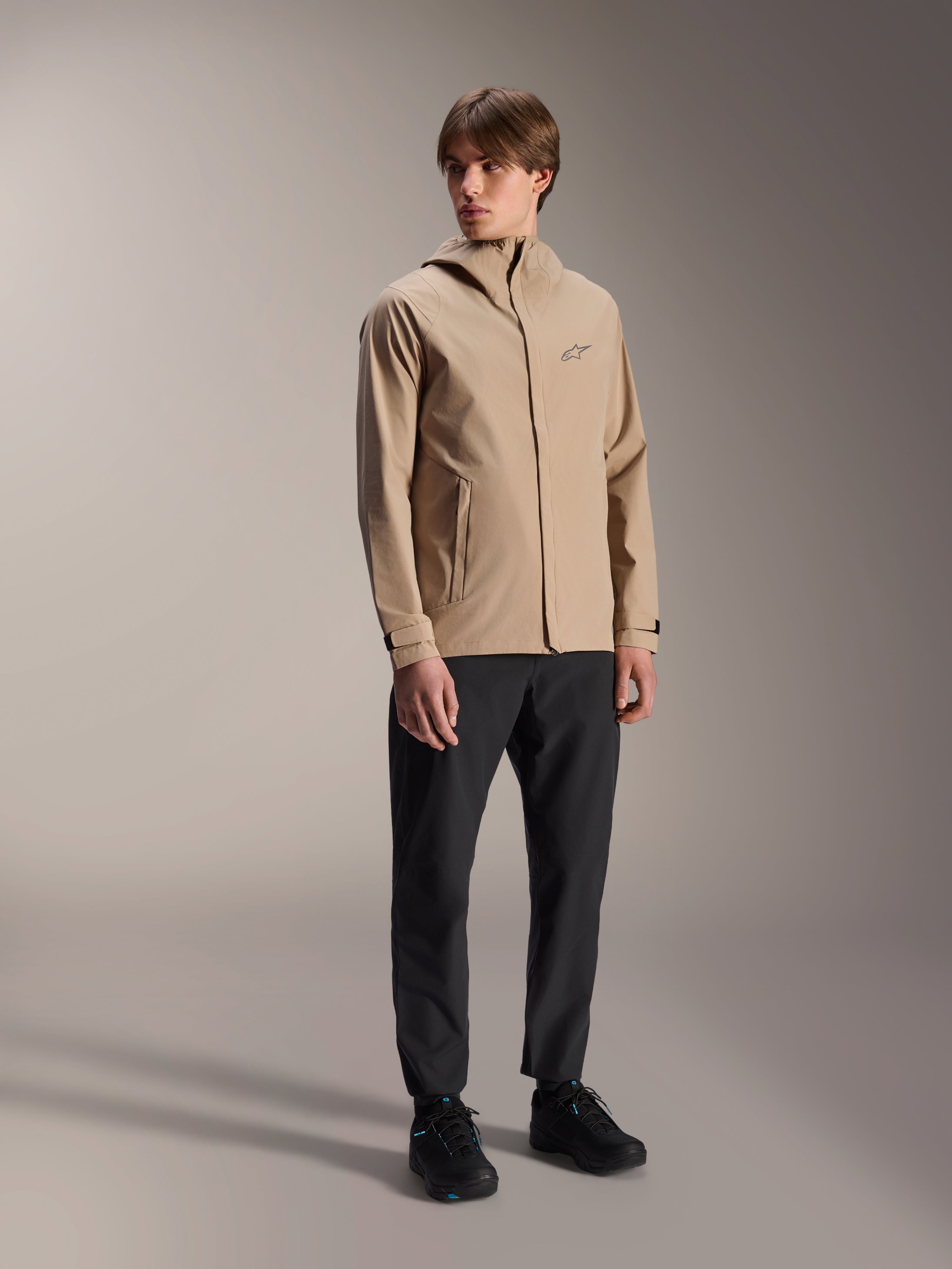 Modelo masculino con la chaqueta de lluvia Alpinestars A-Dura en color marrón claro, combinada con pantalones y calzado negros, chaqueta impermeable minimalista para ciclismo de montaña con diseño de capucha y un pequeño logotipo en el pecho