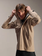 Alpinestars A-Dura Rain Jacket, chaqueta de ciclismo MTB, marrón claro, modelo vistiendo la chaqueta y ajustándose la capucha elástica, con logotipo negro en el pecho, bolsillos con cremallera y tejido técnico impermeable de 2.5 capas