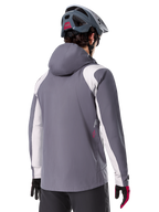 Chaqueta impermeable Alpinestars A-Dura, chaqueta de mountain bike, gris titanio oscuro, vista trasera con paneles en gris claro en hombros y brazos, detalle de logotipo rojo en el lateral, usada con casco de MTB gris y guantes negros y rojos, ropa técnica para ciclismo de montaña