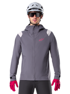 Alpinestars A-Dura Rain Jacket, chaqueta impermeable para ciclismo de montaña, gris titanio oscuro, diseño con capucha con logotipo Alpinestars rosa y paneles gris claro en los hombros, usada con casco de MTB blanco y guantes rosa brillante, equipamiento impermeable para ciclismo todoterreno
