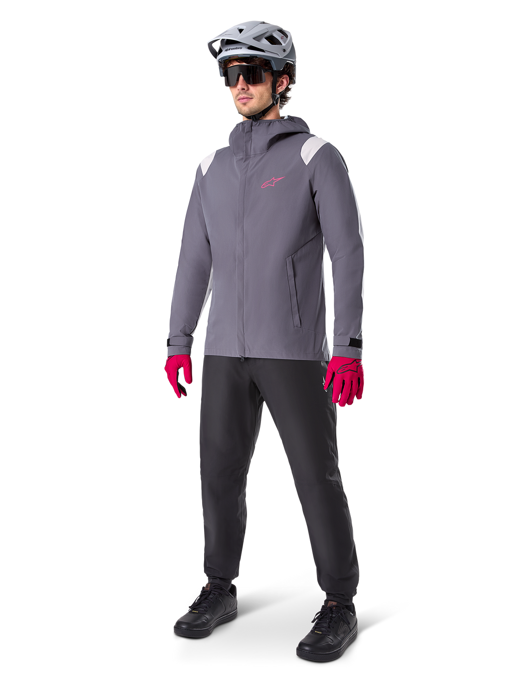Modelo masculino con la chaqueta impermeable Alpinestars A-Dura en color Dark Titanium, chaqueta gris con capucha, logotipo rosa en el pecho y paneles gris claro en los hombros, combinada con casco de MTB gris, gafas de sol oscuras, guantes rosa vibrante y pantalones negros para ciclismo de montaña.