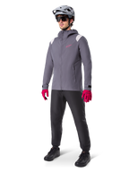 Modelo masculino con la chaqueta impermeable Alpinestars A-Dura en color Dark Titanium, chaqueta gris con capucha, logotipo rosa en el pecho y paneles gris claro en los hombros, combinada con casco de MTB gris, gafas de sol oscuras, guantes rosa vibrante y pantalones negros para ciclismo de montaña.